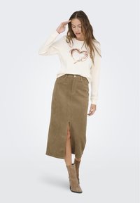 Suéter crema con un diseño de corazón metálico, combinado con una falda de ante color caqui con abertura frontal y botas de ante marrón hasta los tobillos.