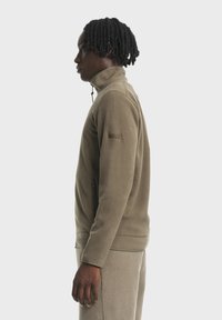 Fleecejacke in Olivgrün mit hohem Kragen, halbem Reißverschluss und seitlicher Reißverschlusstasche; verfügt über eine strukturierte Oberfläche und ein Logo-Patch.