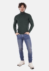 Maglione verde a collo alto dal fitting aderente, abbinato a jeans azzurri chiari e sneakers bianche. Il maglione presenta una texture liscia e orlo a coste.