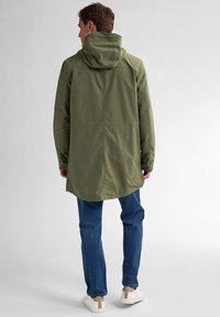 Olijfgroene waterdichte jas met een hoge kraag en capuchon, lange mouwen en een relaxte pasvorm, gecombineerd met blauwe jeans en witte sneakers.