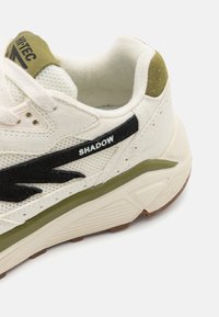 Hvit sneaker med svarte og olivengrønne detaljer, med et teksturert mesh-overdel, polstret krage og en gummigummisåle. "SHADOW"-merket synlig.