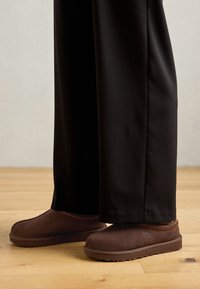 Brown suede slippers with a rubber sole, featuring orange stitching along the collar, paired with black pants on a wooden floor.