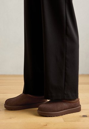 Chaussons en daim marron avec une semelle en caoutchouc, ornés de surpiqures orange le long du col, associés à un pantalon noir sur un sol en bois.