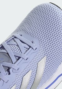 Chaussure de sport légère en violet clair avec tige en mesh, embout renforcé, accents gris, rayures argentées et lacets texturés sur une semelle blanche.