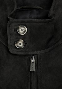 Kołnierz czarnej zamszowej kurtki z dwoma przezroczystymi guzikami i metalowym suwakiem z wygrawerowanym napisem "Scalpers."