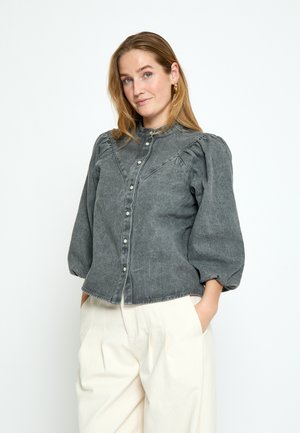Vrouw met lichtbruin haar, gekleed in een grijze blouse met knopen en opgezette mouwen, en witte broek met wijde pijpen, handen in de zakken, neutrale achtergrond.