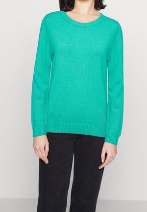 Femme portant un pull turquoise uni à manches longues et un pantalon noir, debout devant un fond clair uni.