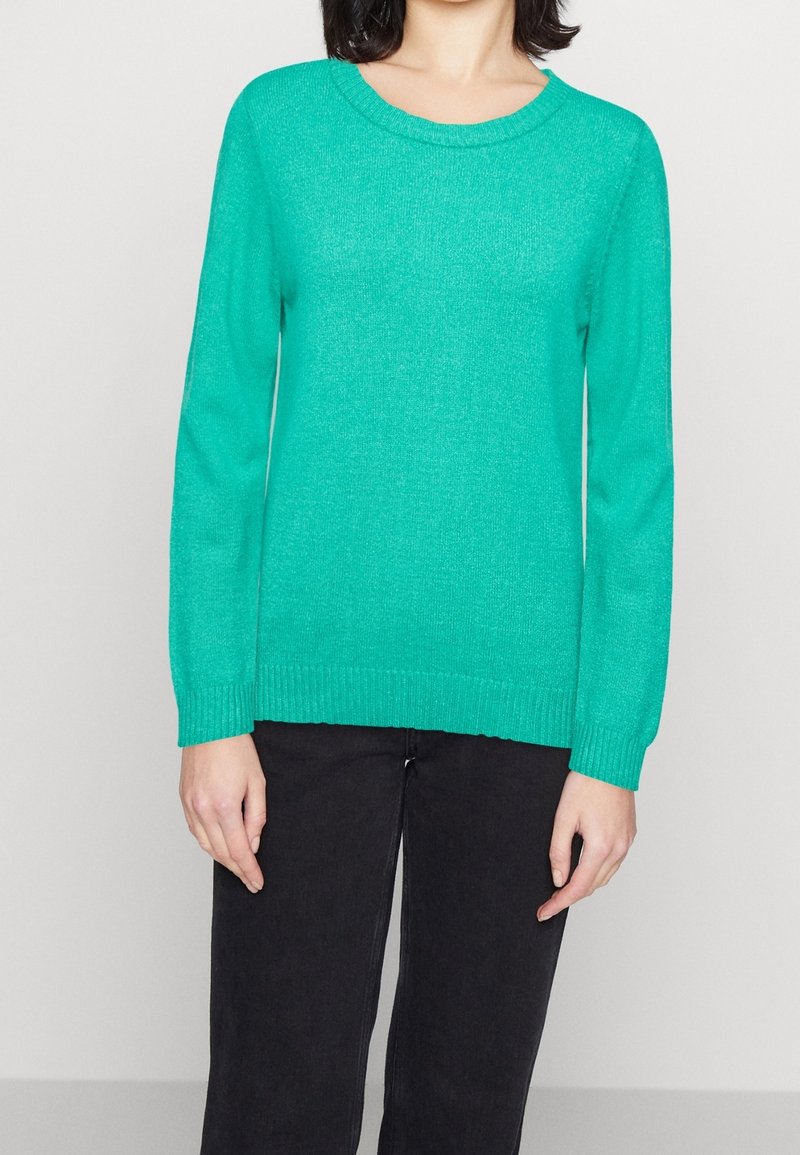 Femme portant un pull turquoise uni à manches longues et un pantalon noir, debout devant un fond clair uni.