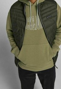 Gilet verde trapuntato indossato sopra un cappuccio oliva abbinato con tasca frontale. Entrambi presentano dei lacci; i jeans scuri completano l'outfit.