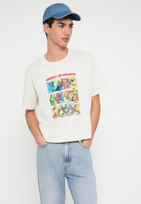 Majica krem boje s šarenim grafičkim prikazom strip panela DC superheroja. Nošena s svijetloplavim trapericama i plavom kapom.