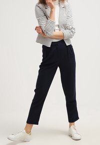 Pantalon bleu marine à coupe décontractée avec plis à l'avant, associé à un blazer blanc et rayé bleu marine, et des baskets blanches avec des accents jaunes.