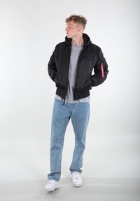 Black bomber jacket with a hood, ribbed cuffs, and a red tag on the sleeve, paired with light blue jeans and white sneakers.