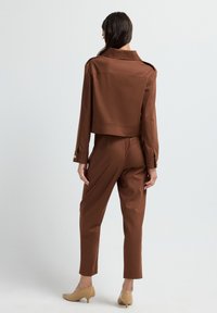 Kvinna sedd bakifrån med en brun långärmad kort jacka och matchande byxor, samt beiga kitten heel-skor, stående på vit bakgrund.