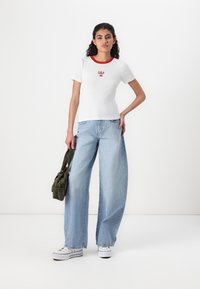 White short-sleeve T-shirt with red trim and GAP logo, paired with light blue wide-leg jeans and white sneakers, holding a green bag.