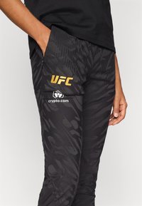Svarte treningsbukser med et strukturert mønster, med en gull "UFC" logo og en hvit "crypto.com" grafikk på sidelommen.