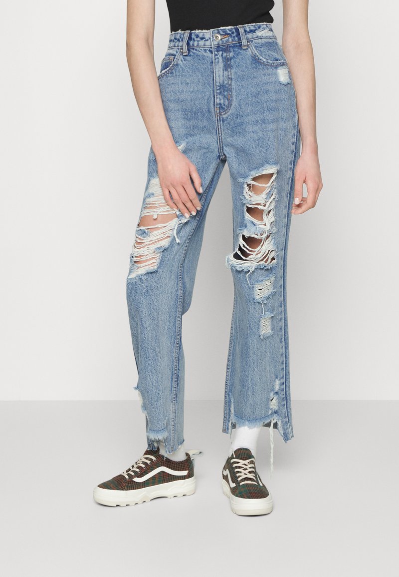 High-waisted blue denim jeans featuring multiple rips and frayed hems. Paired with plaid sneakers, presenting a casual look.