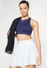 Top corto deportivo de color marino con un acabado texturizado, combinado con una falda blanca de corte acampanado. La modelo sostiene una toalla negra, mostrando el diseño de ropa activa.