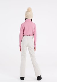 Svetlorožový top s dlhým rukávom, krémové lyžiarske nohavice a béžová pletená čiapka s pom-pomom. Čierne topánky dotvárajú outfit.