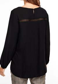 Blusa negra con cuello redondo, espalda plisada y detalles de yugo transparente. Mangas largas con puños abotonados. Tejido suave y ligero.