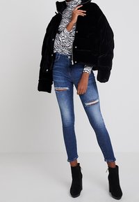 Chaqueta acolchada negra, camiseta de manga larga con estampado de cebra, jeans ajustados desgastados y botines negros con detalles dorados.
