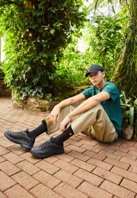 Black sneakers with textured upper, grey accents, and grey laces. Model wears khaki pants, black socks, and a green t-shirt in a lush setting.