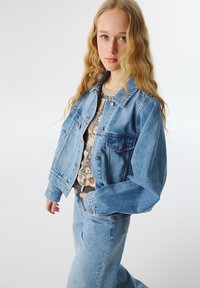 Chaqueta de mezclilla azul con un corte relajado, que presenta dos bolsillos en el pecho y un cierre de botones, combinada con una blusa beige de estampado y unos vaqueros a juego.
