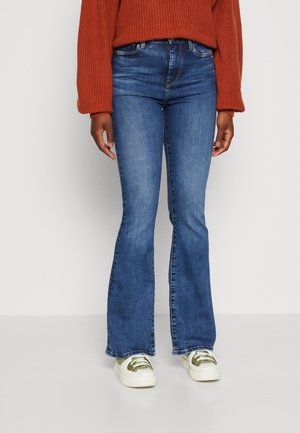 Person iført blå vidde jeans, hvide sneakers med olivengrønne detaljer og en orange strikket sweater, stående mod en ensfarvet hvid baggrund.