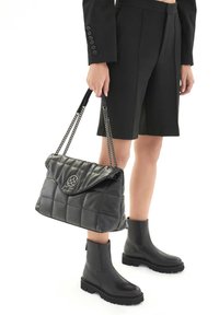 Borsa a tracolla nera trapuntata con manico a catena, dettaglio logo in argento e finitura lucida; abbinata a pantaloni corti neri e stivali alla caviglia.