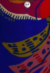 Pañuelo de seda con patrones coloridos sobre un fondo azul; incluye un diseño de hoja amarilla y acentos en rosa y verde. Detalle de un pequeño botón visible.