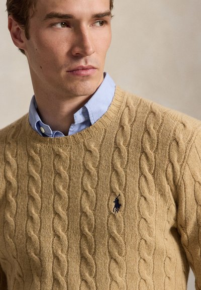 Hombre con camisa azul claro con cuello bajo un suéter de punto trenzado color tostado con logo de polo azul marino, mirando de lado sobre fondo beige.