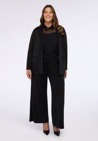 Zwarte op maat gemaakte blazer met een top met pailletten kraag, gecombineerd met zwarte wijde broek. Gladde stof met subtiele glans, afgewerkt met zwarte hakken.