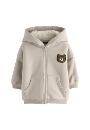 Bež kapuutsiga jakk, mille sees on pehme fleece'iga vooder. Ees on karupatch ja kaks eesmist taskut. Lisatud on soonilised varrukad.