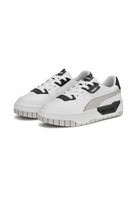 Paire de baskets blanches Puma avec des accents noirs et gris, semelles épaisses et lacets, présentées légèrement en angle sur un fond blanc.