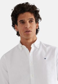 Camicia bianca con colletto, dotata di un piccolo logo blu sul petto sinistro. Realizzata in cotone con una texture liscia.