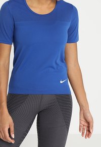 Camiseta deportiva azul de Nike con mangas cortas, diseño de malla en los hombros, cuello redondo y logo blanco en la parte inferior izquierda. Combinada con leggings negros con estampado.