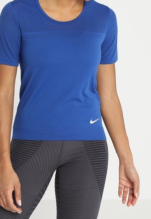 Camiseta deportiva azul de Nike con mangas cortas, diseño de malla en los hombros, cuello redondo y logo blanco en la parte inferior izquierda. Combinada con leggings negros con estampado.