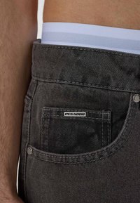 Sive traperice s ravnim krojem i vidljivom stražnjom džepom s metalnom oznakom marke. Pojas uključuje bijelu elastičnu traku.