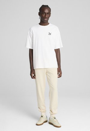 White cotton t-shirt with short sleeves and a black logo on the chest, paired with cream-colored sweatpants and beige sneakers.