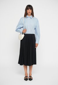 Camicia a righe blu e bianche con maniche lunghe, abbinata a una gonna plissettata nera e ballerine nere; tiene una piccola borsa bianca.
