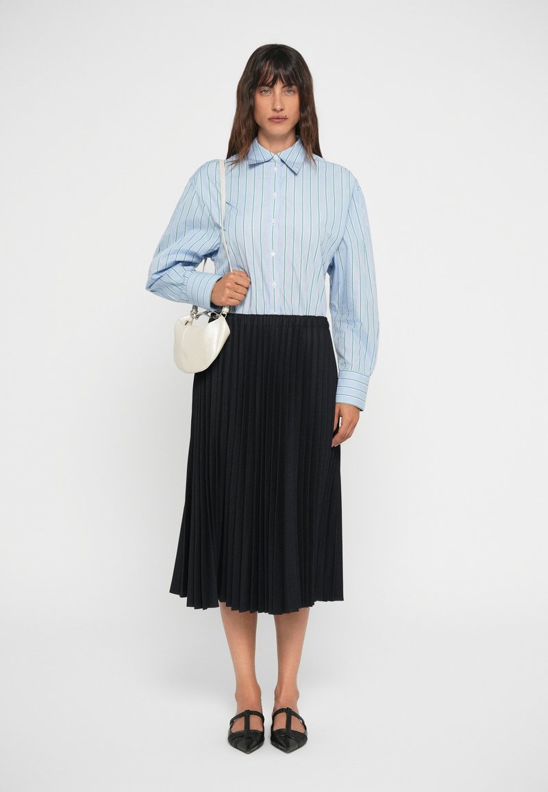 Camicia a righe blu e bianche con maniche lunghe, abbinata a una gonna plissettata nera e ballerine nere; tiene una piccola borsa bianca.