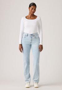 Γυναίκα όρθια που φοράει λευκό μπλουζάκι μακρυμάνικο, ανοιχτόχρωμα τζιν ψηλόμεσα, λευκά αθλητικά παπούτσια, με σκουλαρίκια και δαχτυλίδια.