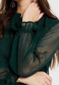 Blusa verde scuro con maniche a sbuffo trasparenti, scollatura a volant e una sottile trama. I polsini arricciati aumentano la morbidezza e l'eleganza.