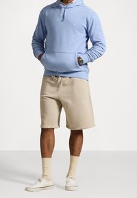 Man wearing light blue hoodie, beige shorts, beige crew socks, white sneakers, and a wristwatch, standing with hands in hoodie pockets.