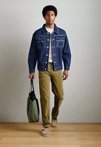 Jeansjakke med to brystlommer, mørkeblå farve, parret med en lys beige t-shirt og olivengrønne bukser, som holder en grøn taske.