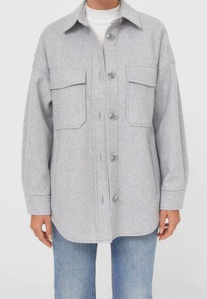 Lichtgrijs wollen jasje met knopen, twee borstflapzakken en zijsteekzakken, gedragen over een witte coltrui en blauwe jeans.