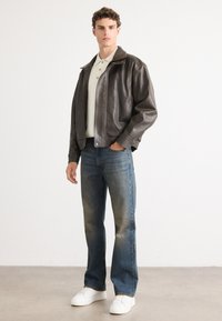 Chaqueta bomber de piel marrón con cierre de cremallera, camiseta polo crema, jeans rectos azul desvanecido y zapatillas blancas. Textura suave.