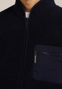 Marinblå fleecejacka med dragkedja fram och ficka på vänster bröst. Mjuk textur, kontrasterande tygficka, minimalistiska detaljer med hårdvara.