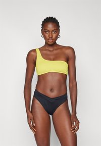 En-skulder bikinitopp i teksturert bright gul med en tettsittende design, kombinert med svarte høyvinklede bikiniunderdeler med en kryssdetalj.