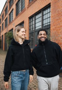 Dvaja jednotlivci nosia čierne fleecové bundy so zipsom a vreckami, v pozadí je tehlová budova. Neformálne džínsy dopĺňajú vzhľad.