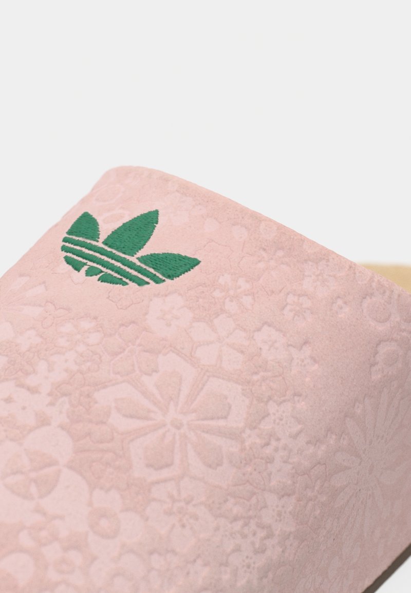 Gros plan sur une surface en daim rose avec des motifs floraux embossés et un logo Adidas vert.